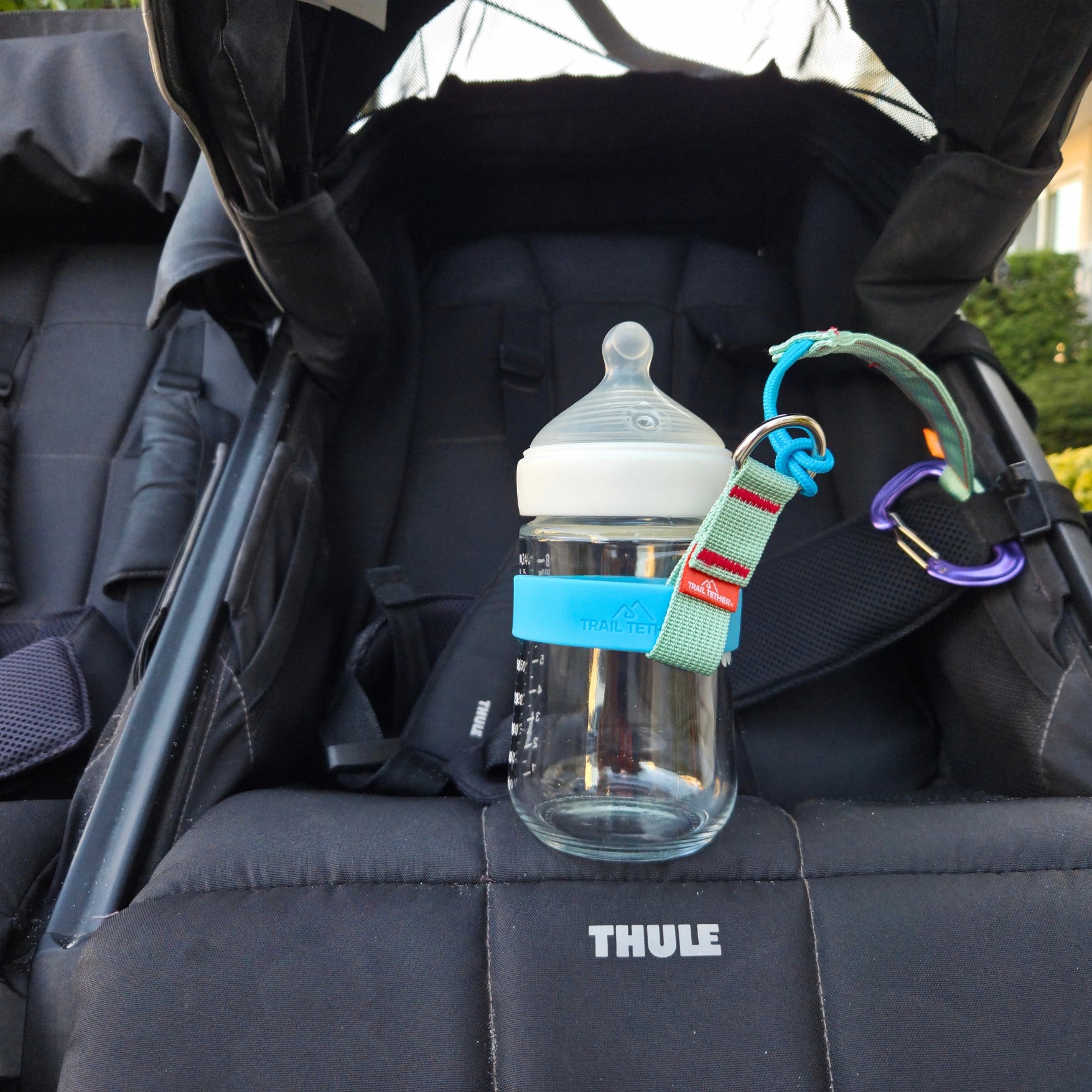A clear baby bottle with a white nipple is clipped to the padded seat of a black Thule stroller. The bottle has a light blue  chameleon ring band and a green-and-blue trail tether strap with a purple carabiner clipping the bottle to the straps, with no child in the seat, shown in an outdoor setting.