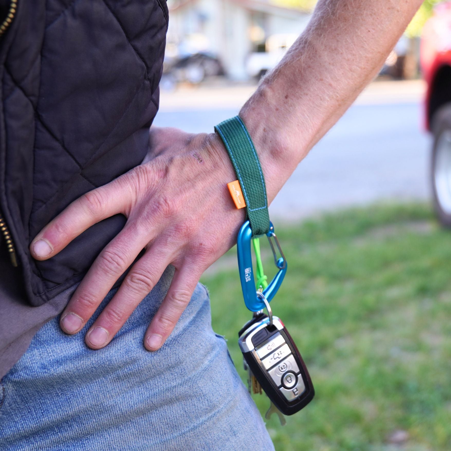 A close-up of a person's hand and waist wearing a trail tether around his wrist, holding a set of keys hands free, including a black car key fob.