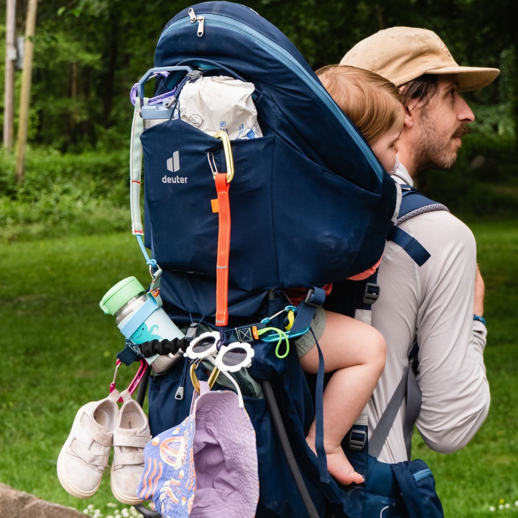 A man outdoors wearing a large navy blue Deuter baby carrier backpack with a child seated in front, plus attached pockets and accessories including shoes, a hat, and a water bottle. tether max is secured many light items to the carrier back.