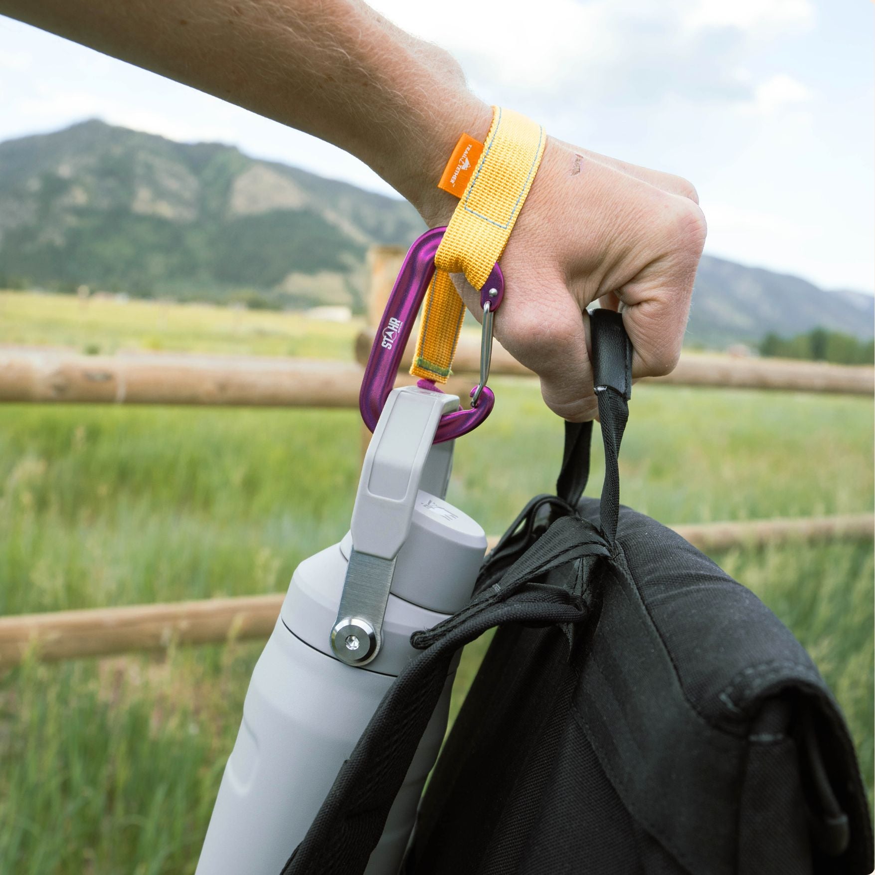 A close-up of a person's hand holding a black backpack and a gray water bottle with a yellow trail tether holding a bottle around the mans wrist, outdoors in a grassy field with mountains in the background.