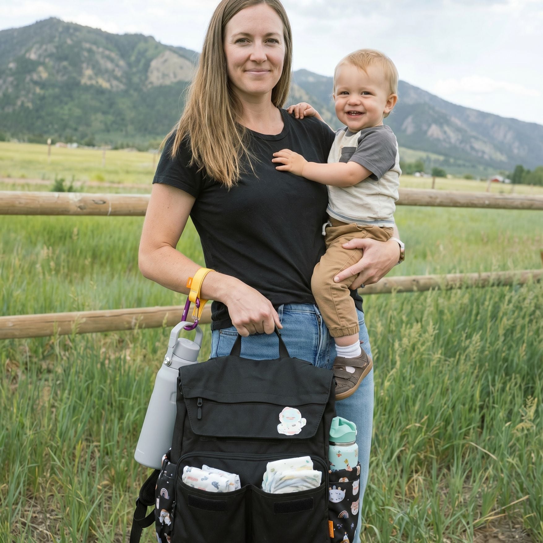A woman outdoors in a grassy field with mountains in the background is holding a baby and wearing a black diaper bag backpack. The diaper bag has multiple pockets containing diapers and accessories, with side pockets holding water bottles. the woman is wearing a bottle bottle around her wrist, held by a yellow trail tether