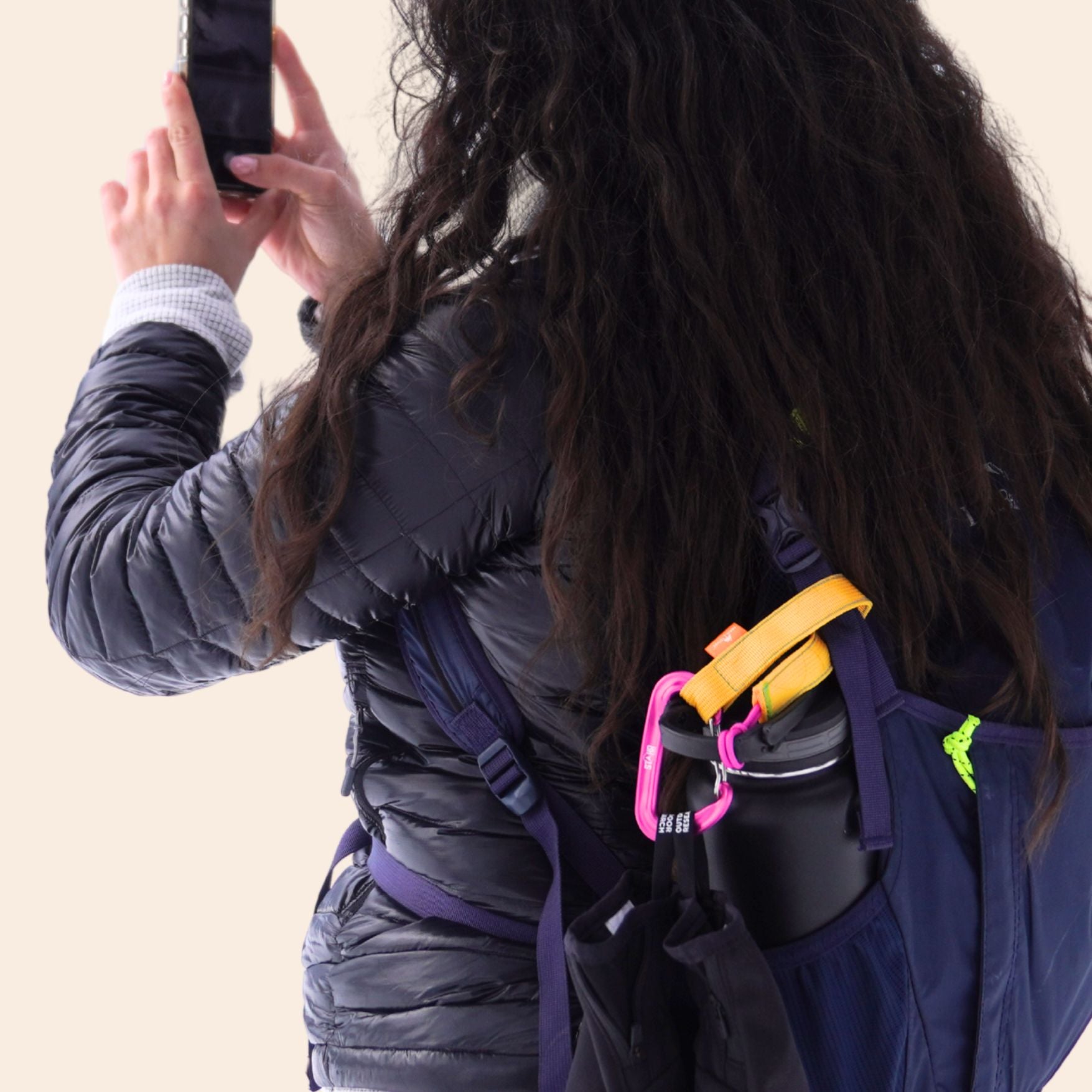 A person wearing a navy blue backpack, shown from behind, holding a smartphone. A black water bottle is secured to the bag by a yellow trail tether, which is also being used to clip a pair of black winter gloves to the backpack