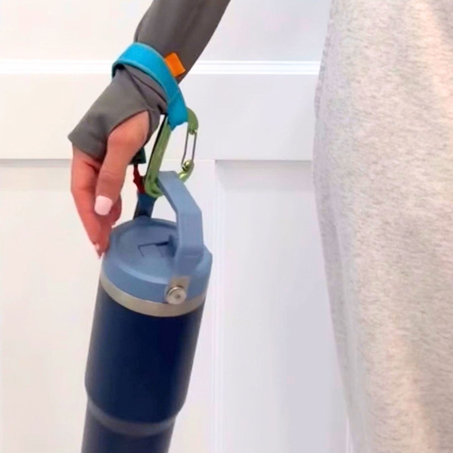 A close-up of a person's hand holding a dark blue insulated bottle from their wrist by using a blue trail tether as a wrist strap to hold the bottle. 