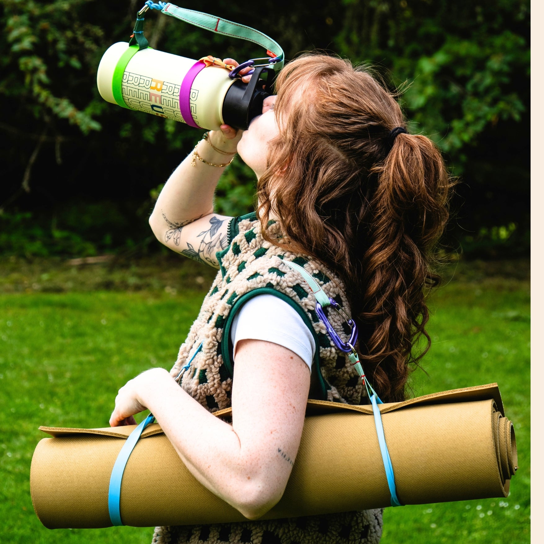 A woman outdoors carrying a rolled tan hiking mat over her shoulder and drinking from a reusable water bottle. the mat has two chameleon ring stretch bands securing it into a roll.  they are connected to a trail tether, which she is using to sling the mat over her shoulder.  the bottle she is drinking from has two chameleon ring silicone bands stretched around the top and bottom.  there is a second trail tether connected to the bottle, which gives it a nice carry handle. 