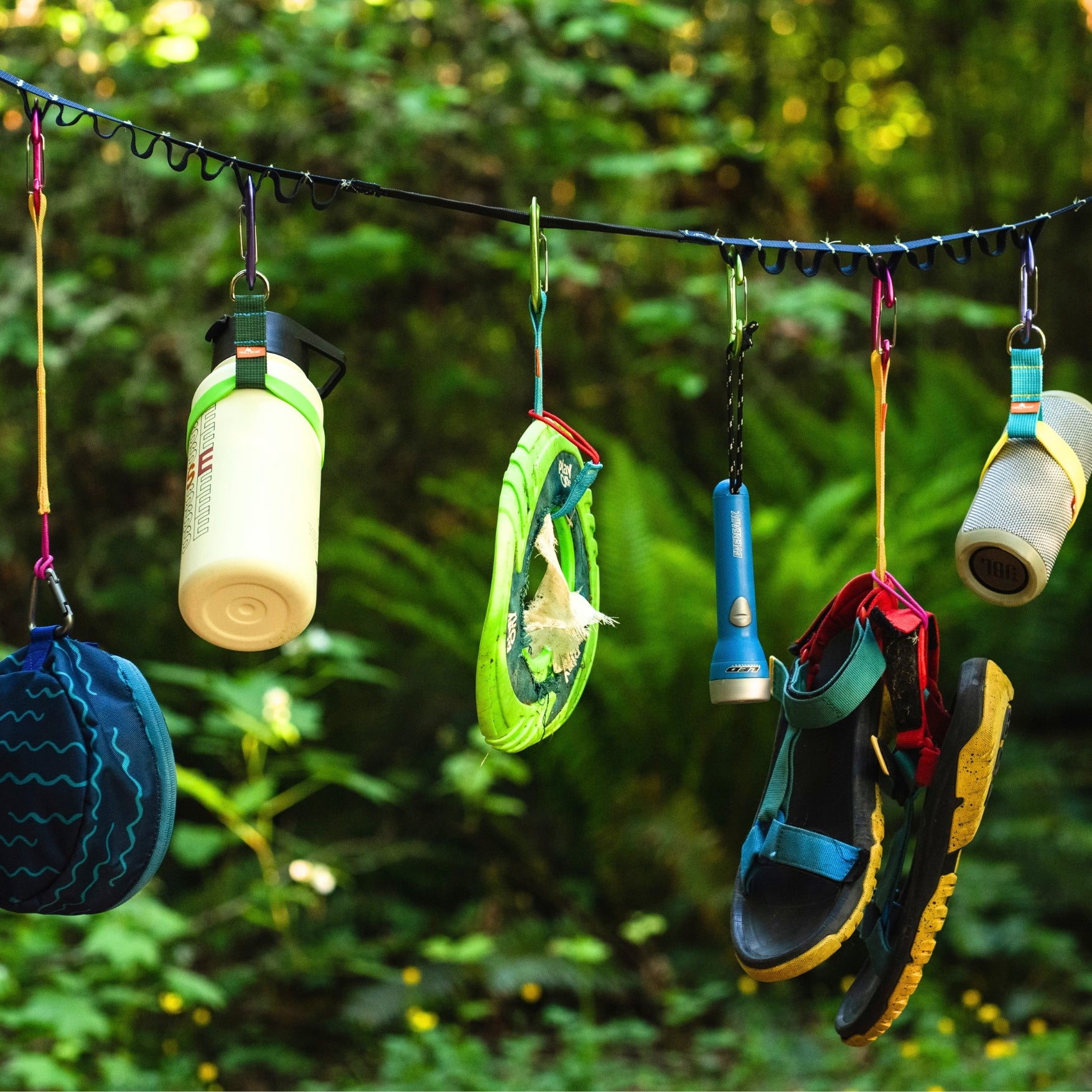 A set of outdoor gear, including water bottles, a sleeping bag, a flip-flop, sandals, and a flashlight, hanging from colorful trail tethers on a tether max strung across a lush green forest background