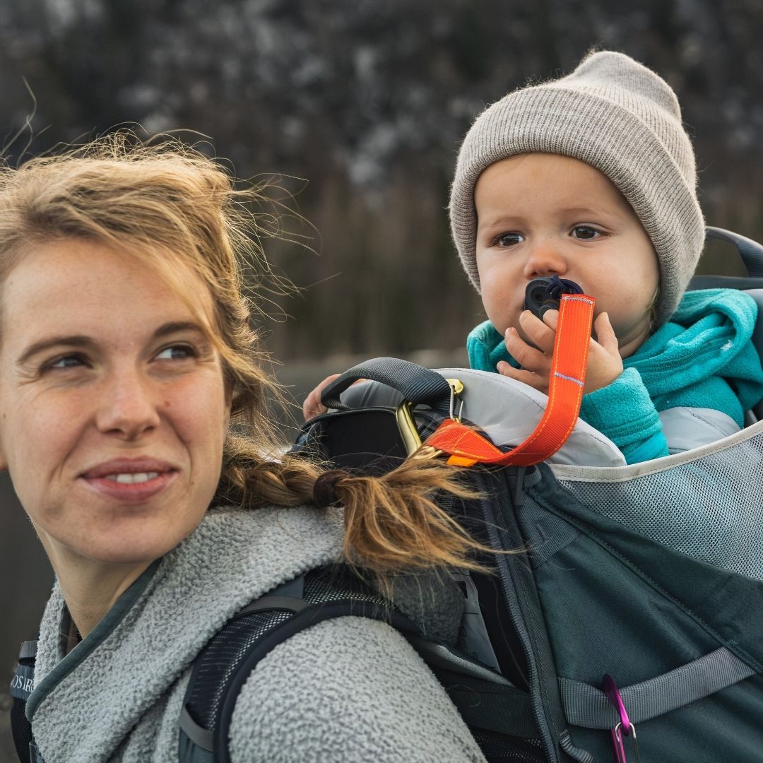 A woman wearing a gray fleece jacket carries a baby in a dark gray backpack carrier outdoors. The baby wears a light gray knit beanie and a turquoise hoodie, with a red trail tether holding a pacifier clip, connected to the carrier