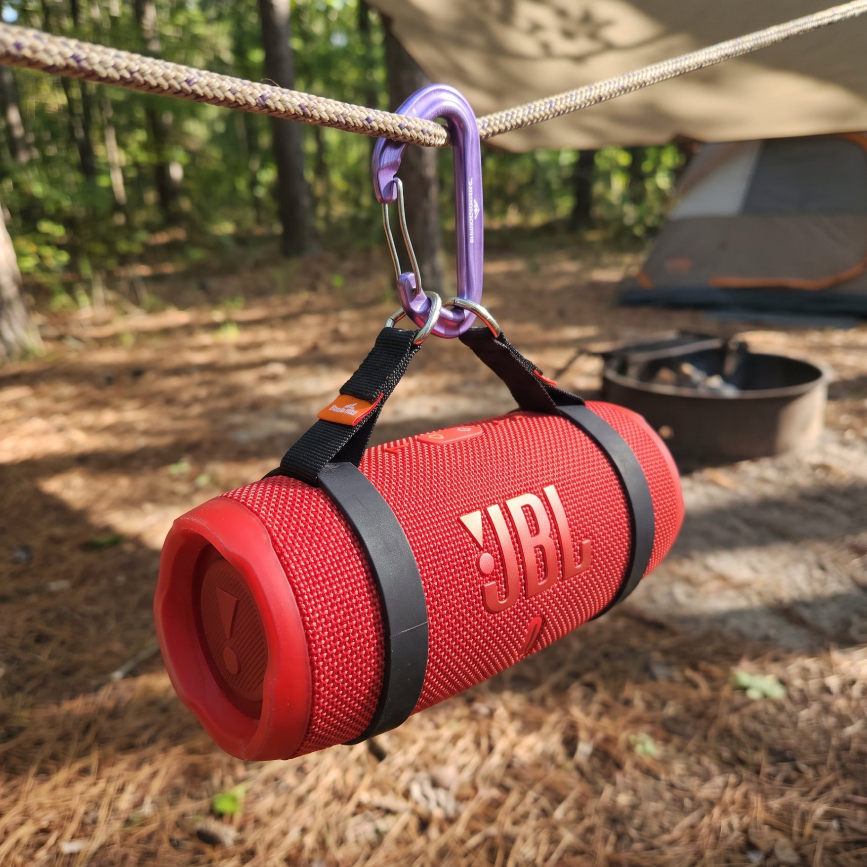 A red JBL portable speaker is secured on either end with two black chameleon ring stretch bands, hanging from a rope in an outdoor campsite setting with a purple carabiner connecting the chameleon rings to the rope, holding the speak suspended.